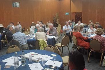 Community meeting in a large room with wooden-panelled walls. Several round tables covered with blue tablecloths are arranged throughout the space, each seating groups of people engaged in discussion. Papers, water bottles, and cups are scattered on the tables, and a stack of chairs is visible against the back wall.