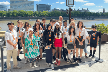 Group of young people and adults standing together outdoors on a sunny day, wearing lanyards with orange badges. They are positioned on a paved terrace overlooking a river, with trees and modern buildings visible across the water. A lamp post and two planters frame the group, and a sign on the left indicates a photo point.