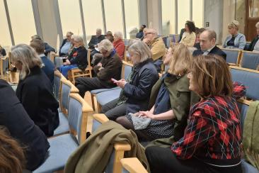 Audience seated in rows of wooden chairs with blue cushions inside a bright room with large frosted windows
