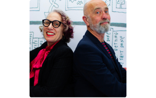 Two adults standing back to back and smiling in front of a whiteboard.