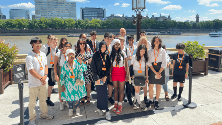 Group of young people and adults standing together outdoors on a sunny day, wearing lanyards with orange badges. They are positioned on a paved terrace overlooking a river, with trees and modern buildings visible across the water. A lamp post and two planters frame the group, and a sign on the left indicates a photo point.