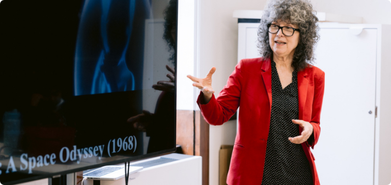 Tutor in a red jacket giving a presentation beside a screen in a classroom