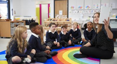 Female teacher holding up a book in front of her class of school kids sitting on the floor in a classroom